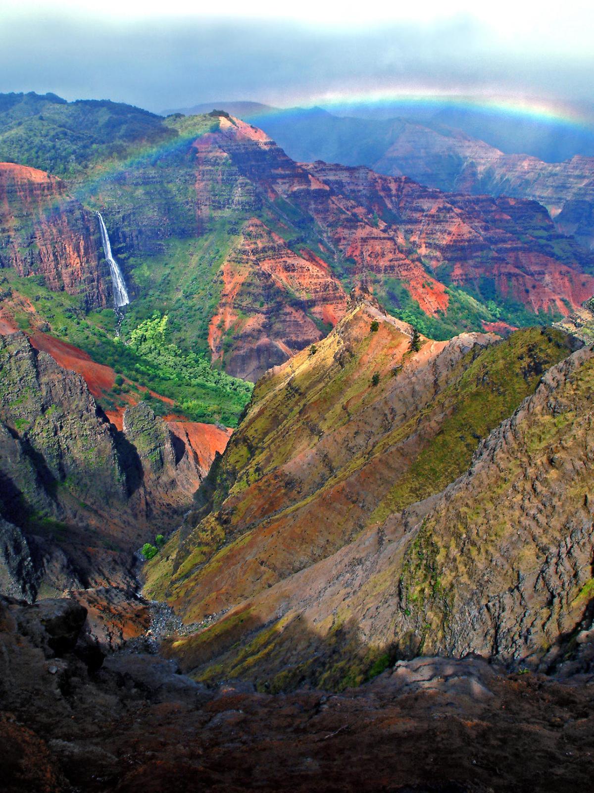 Canyon landscape with waterfall, rainbow, and colorful rock formations under a cloudy sky.