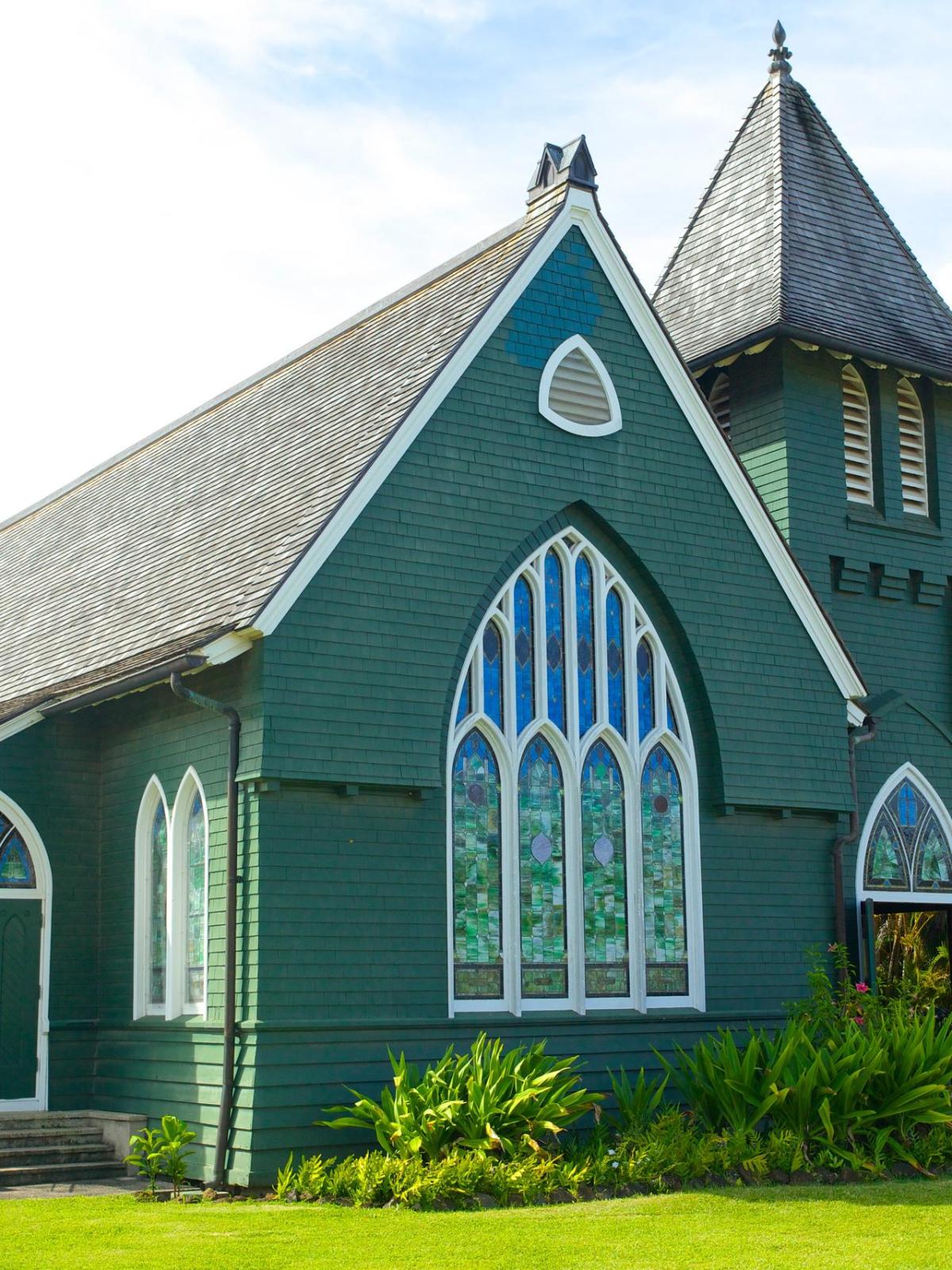 Green wooden chapel with pointed roof and stained glass windows surrounded by tropical plants.