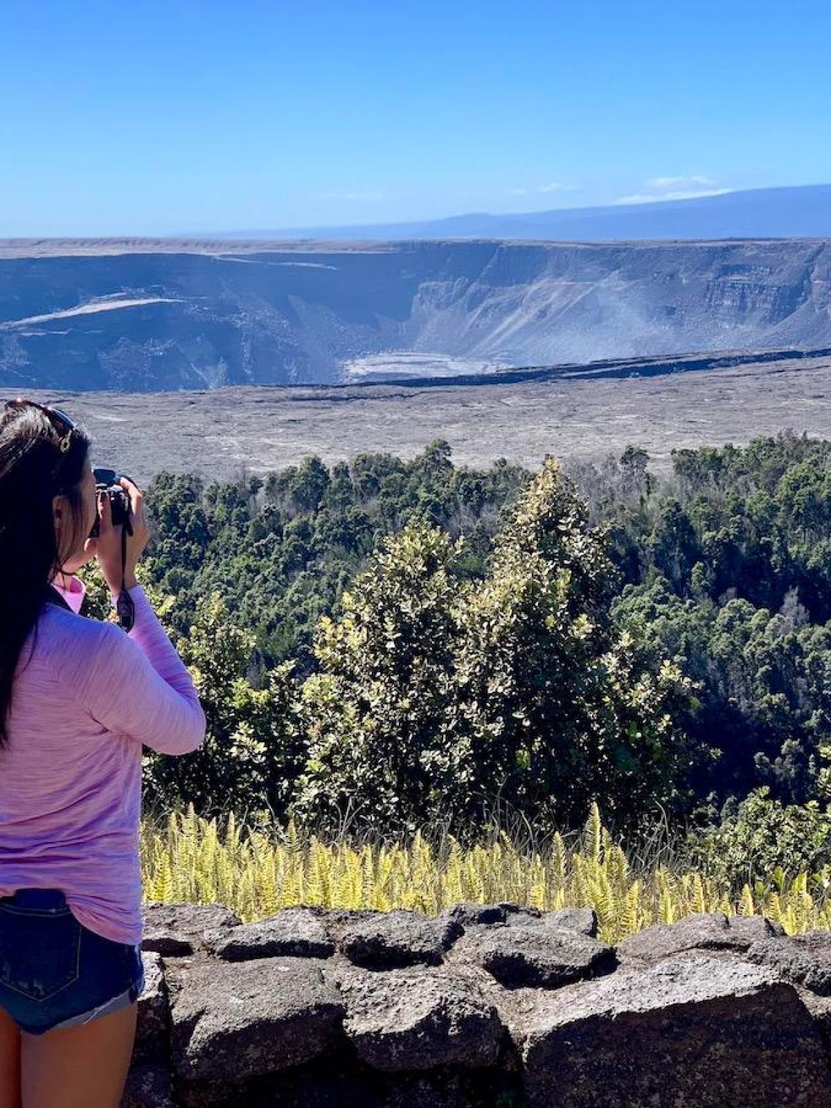 Person photographing a volcanic crater landscape with trees in the foreground.