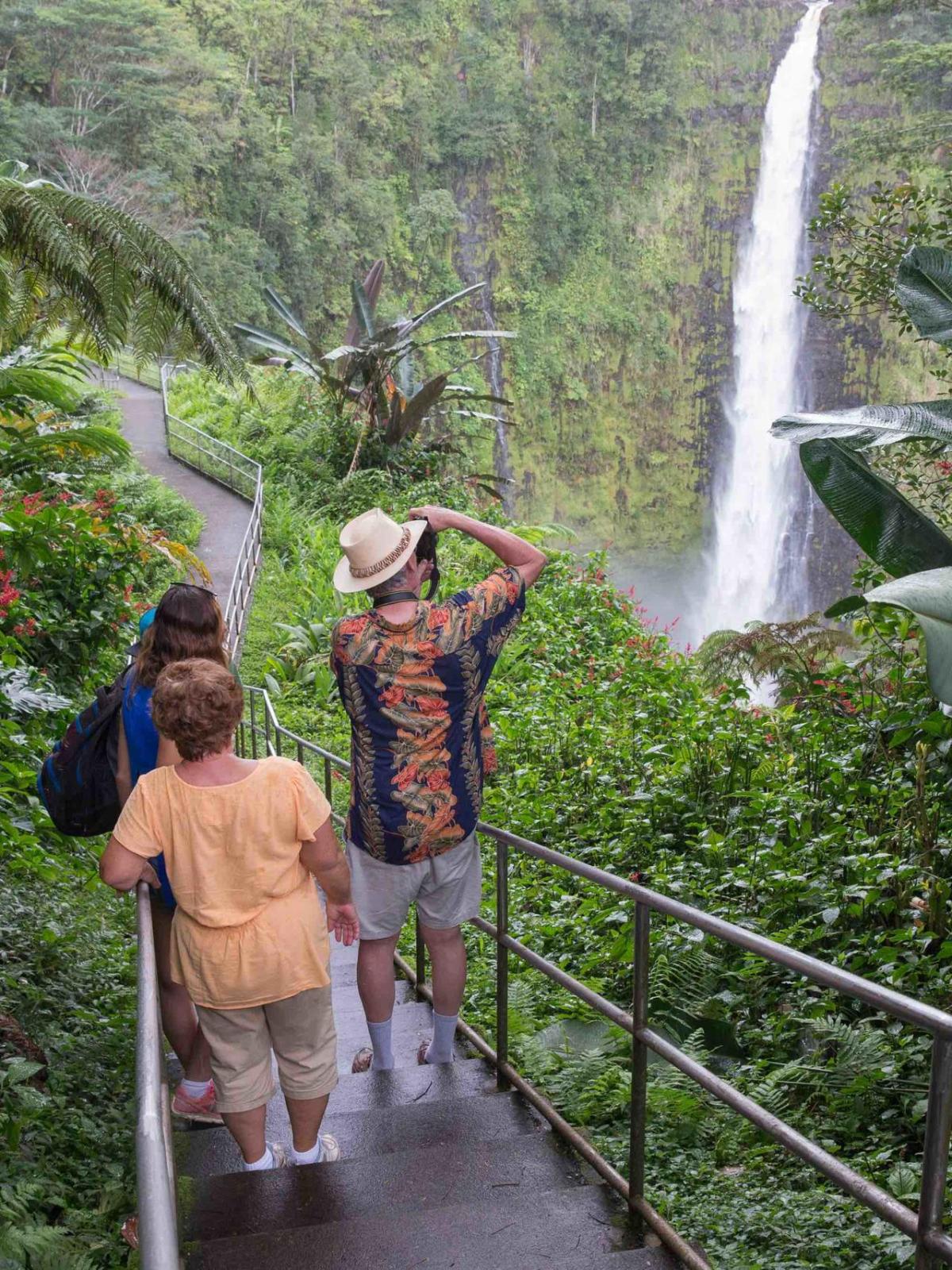 Three people on a path admiring a tall waterfall surrounded by lush greenery.