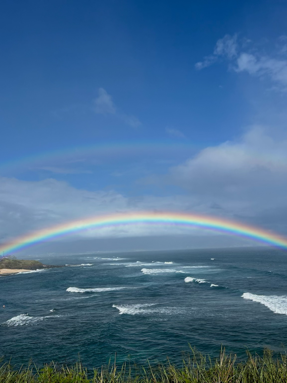 Double rainbow arches over ocean waves under a blue sky and clouds.